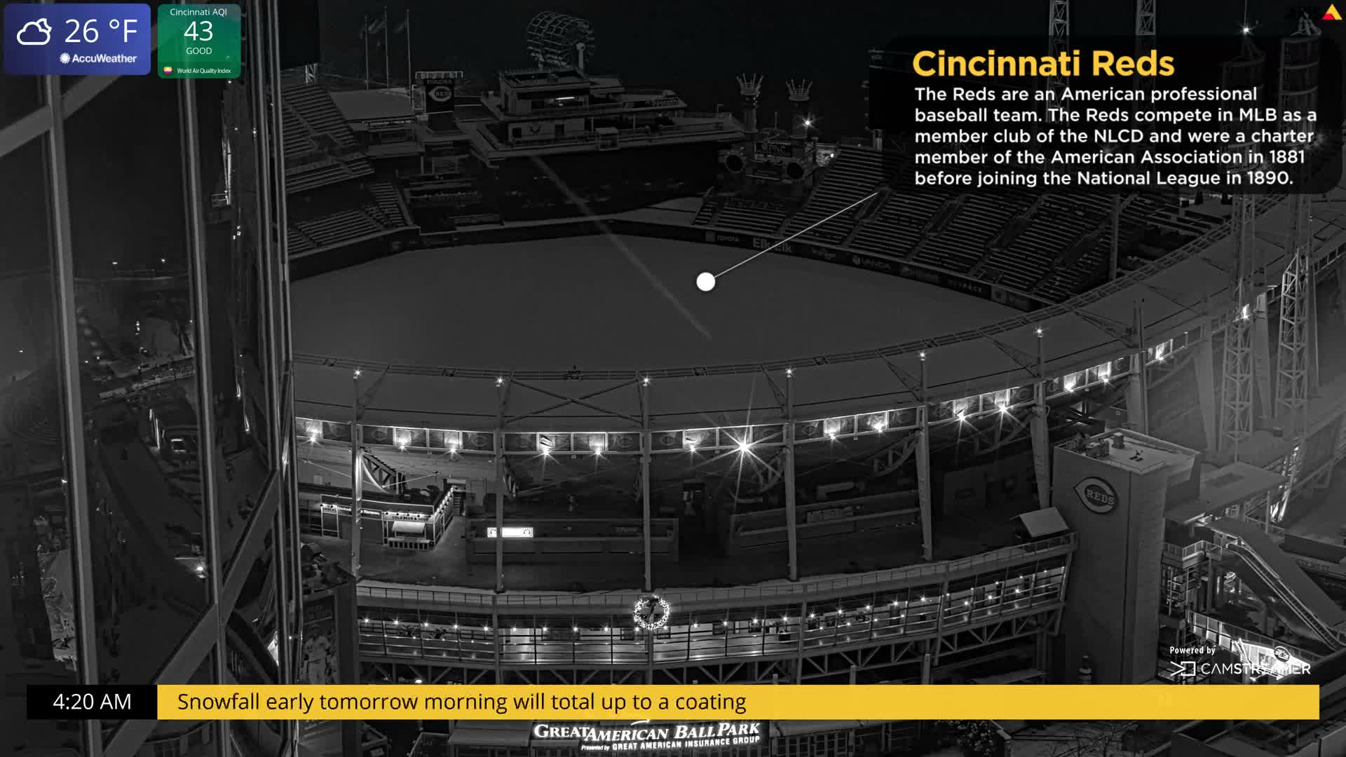 An elevated, monochrome view captures a snow-dusted baseball stadium illuminated by its internal lights on a clear, cold night.