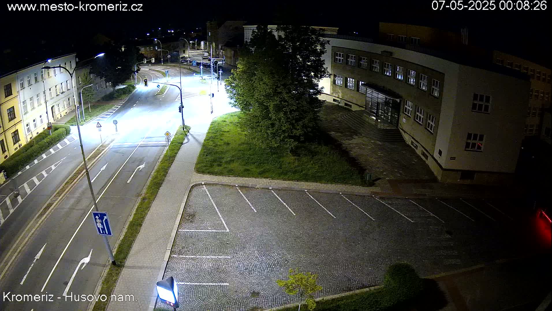 A nighttime view of a mostly empty street and parking lot next to a building, illuminated by streetlights.