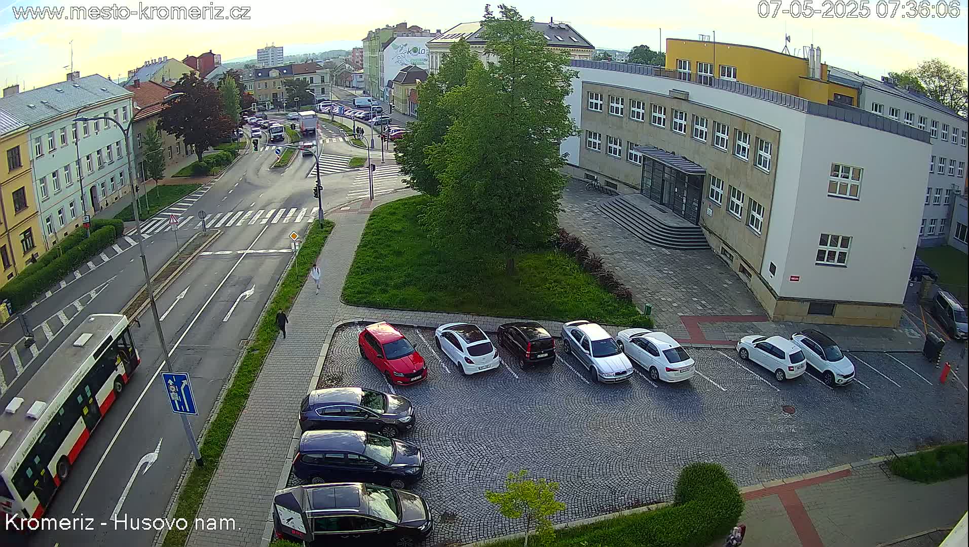 A high-angle, daytime view shows a paved street intersection with parked cars, a bus, pedestrians, and buildings, under a mostly clear sky.