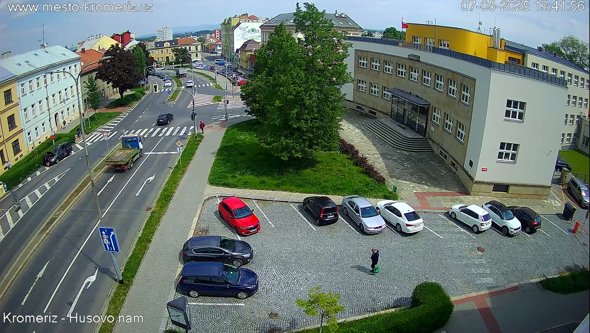 A sunny day reveals a street scene with cars parked in a cobblestone lot next to a building, and several cars driving on a road.