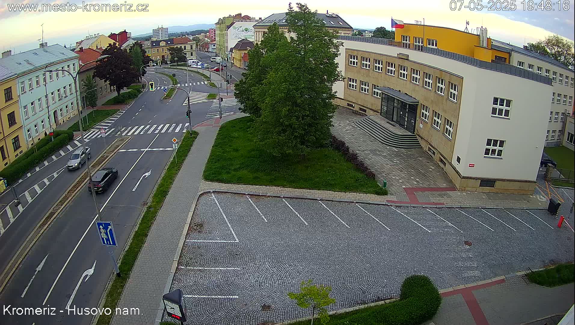 An aerial view of a street intersection with cars, a large building, a paved parking lot, and lush green landscaping under a partly cloudy sky.
