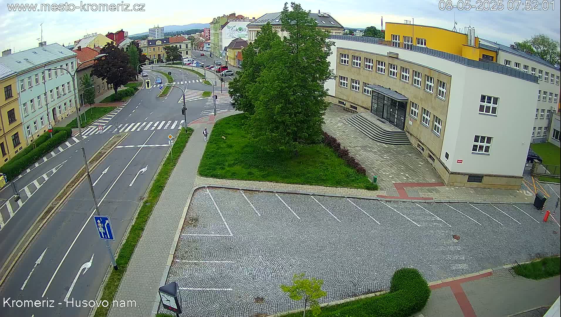 An overcast day reveals a street scene with a large building and an empty cobblestone parking lot, a pedestrian walking on a sidewalk, and several cars parked along the roadside.