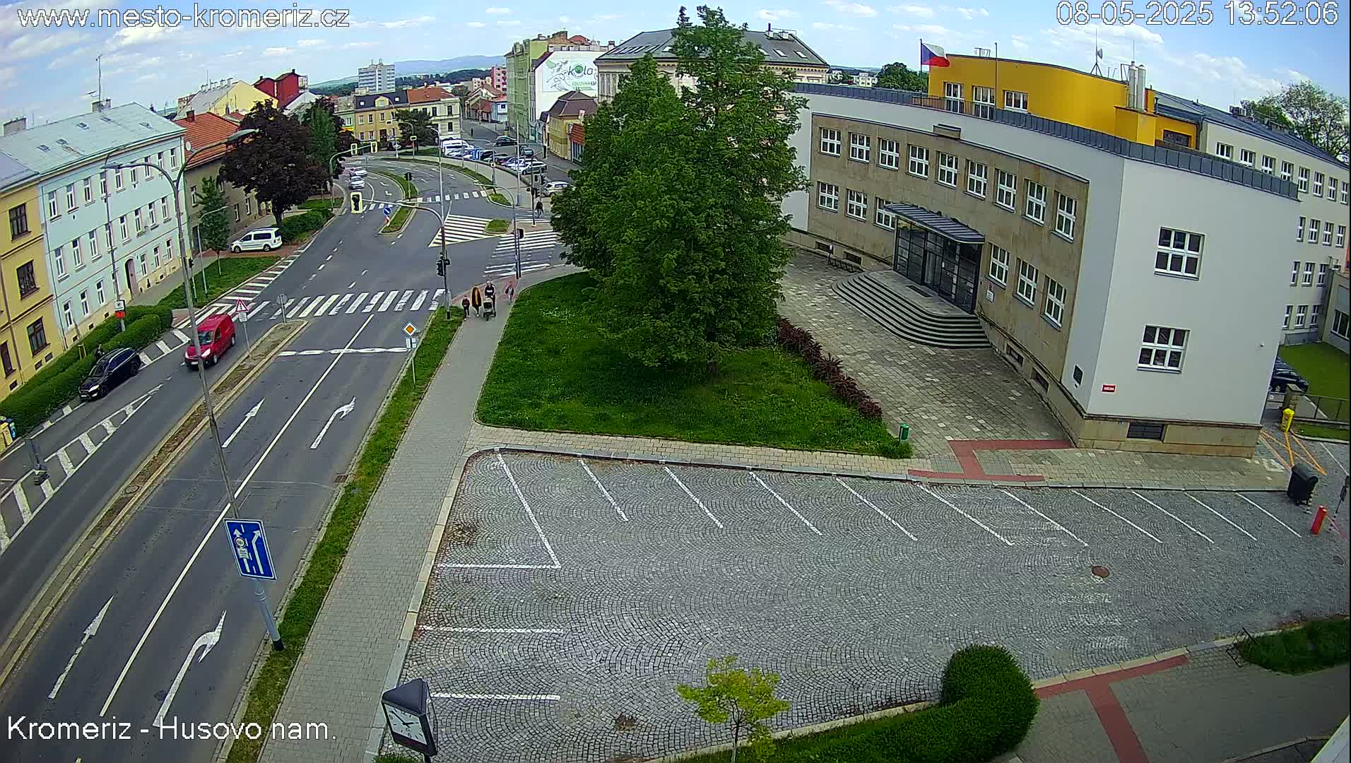 An aerial view shows a city street intersection with cars and pedestrians on a partly cloudy day, next to a large building with a cobblestone parking lot.