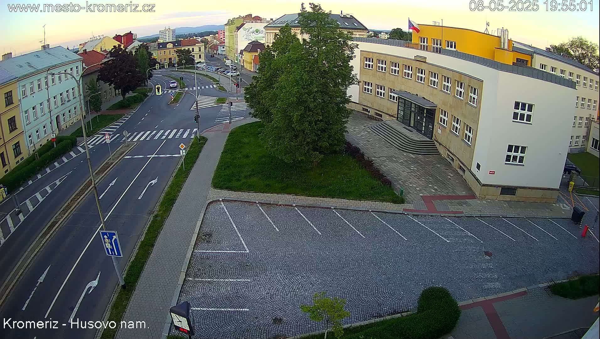 An aerial view shows a street intersection with a cobblestone parking lot and a large building in the evening under a mostly clear sky.