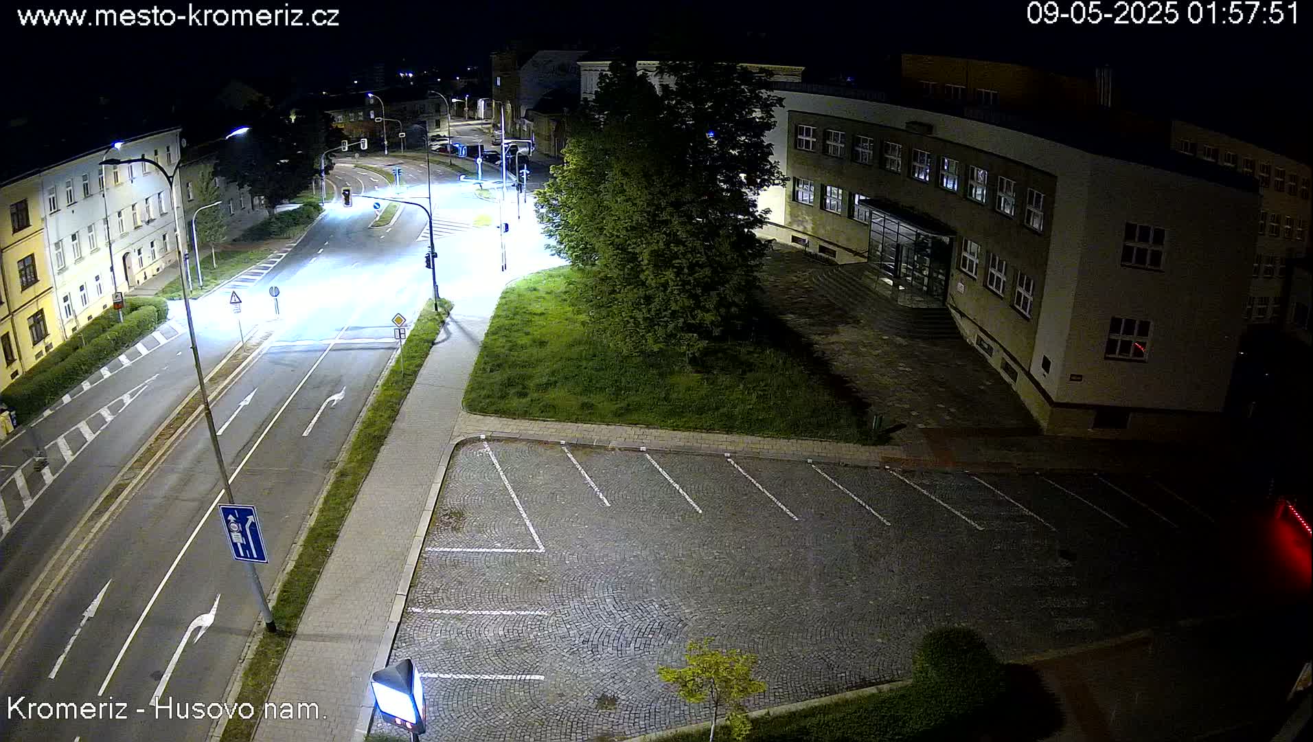 A nighttime, street-level view shows an empty parking lot next to a multi-story building and a two-lane road with minimal traffic.