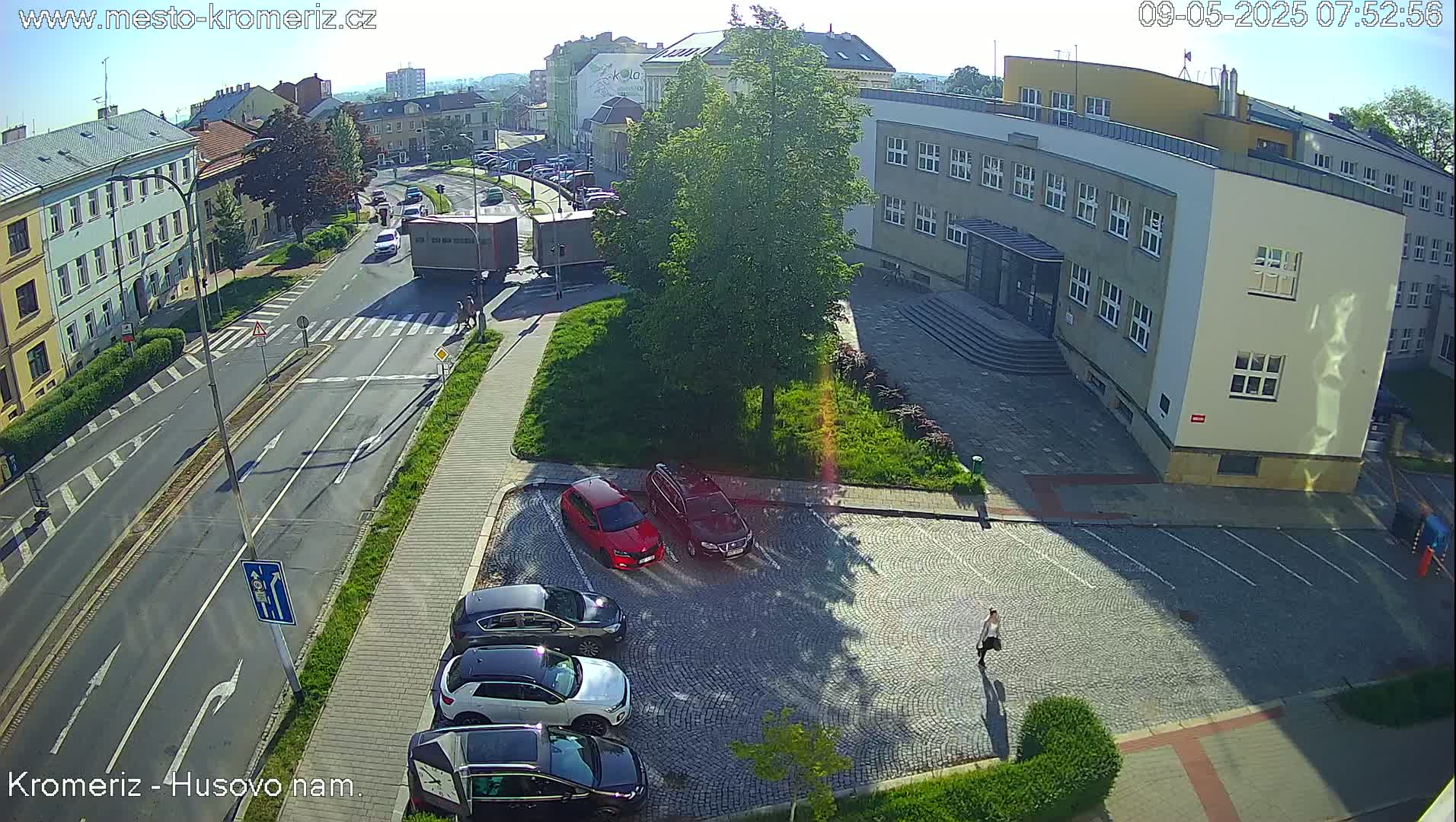 A sunny day view of a town square with cars parked in a cobblestone lot, buildings, and a road with traffic.