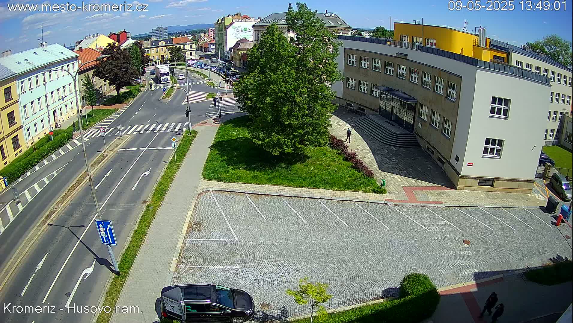A sunny aerial view shows a street intersection with a cobblestone parking lot and a large building nearby.