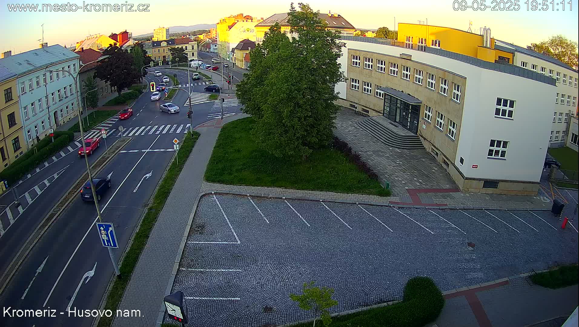 An aerial view of a city street at dusk shows several cars driving on a roadway that intersects with a pedestrian crosswalk, next to a cobblestone parking lot and a large light-colored building.