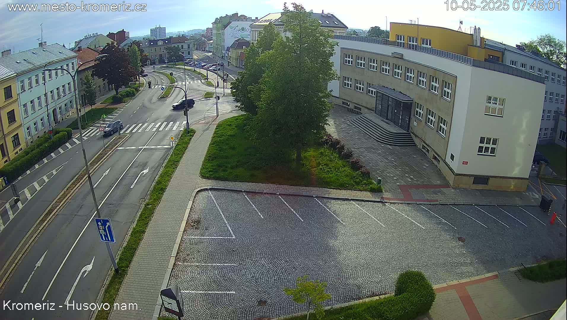 A sunny day reveals a street scene with a cobblestone parking lot next to a large building, and a street intersection with several cars.