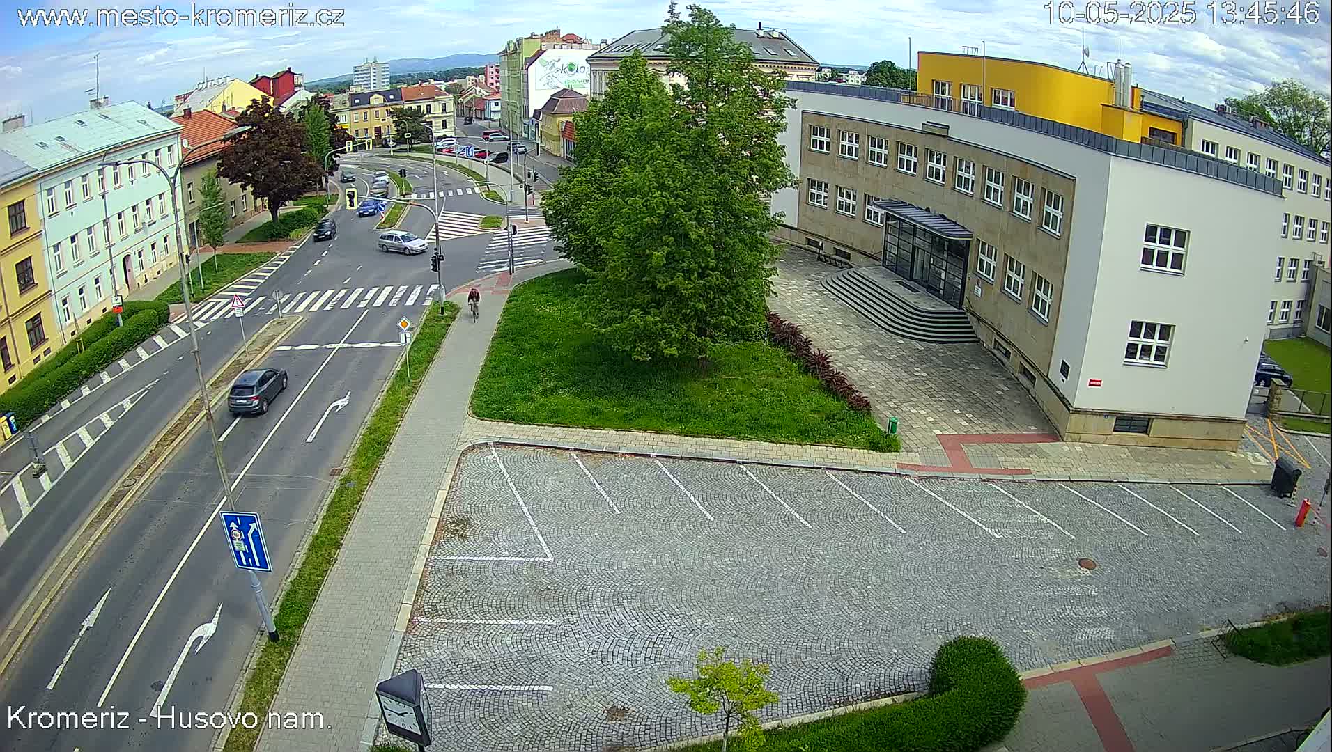 A mostly sunny overhead view of a city street intersection with a large building, a paved parking lot, and a person riding a bicycle on a bike path.