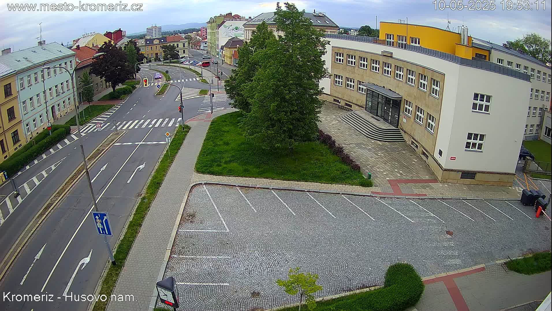 An overhead view shows a mostly empty cobblestone parking lot next to a multi-story building on a partly cloudy day, with a street and pedestrian crosswalks visible in the background.