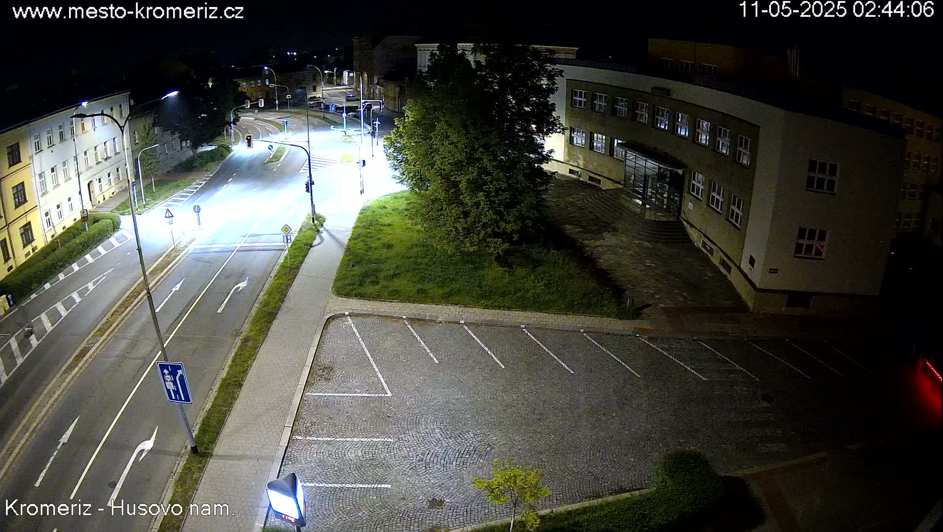 A nighttime aerial view shows a mostly empty street and parking lot next to a building, illuminated by streetlights.