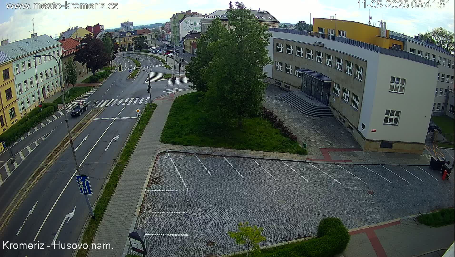 An overcast day shows a street intersection with a building and a cobblestone parking lot, surrounded by green landscaping.
