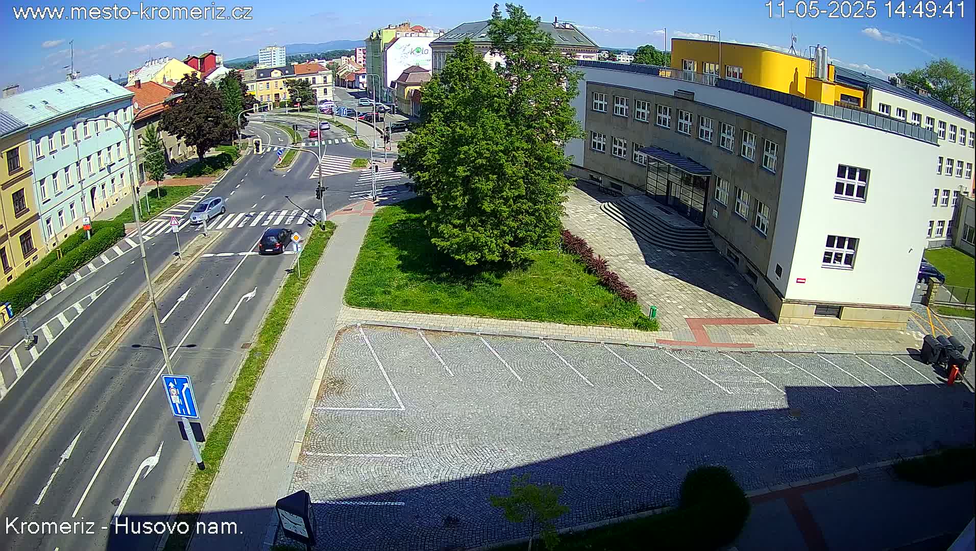 A sunny aerial view shows a street intersection with cars, a parking lot, and a multi-story building.