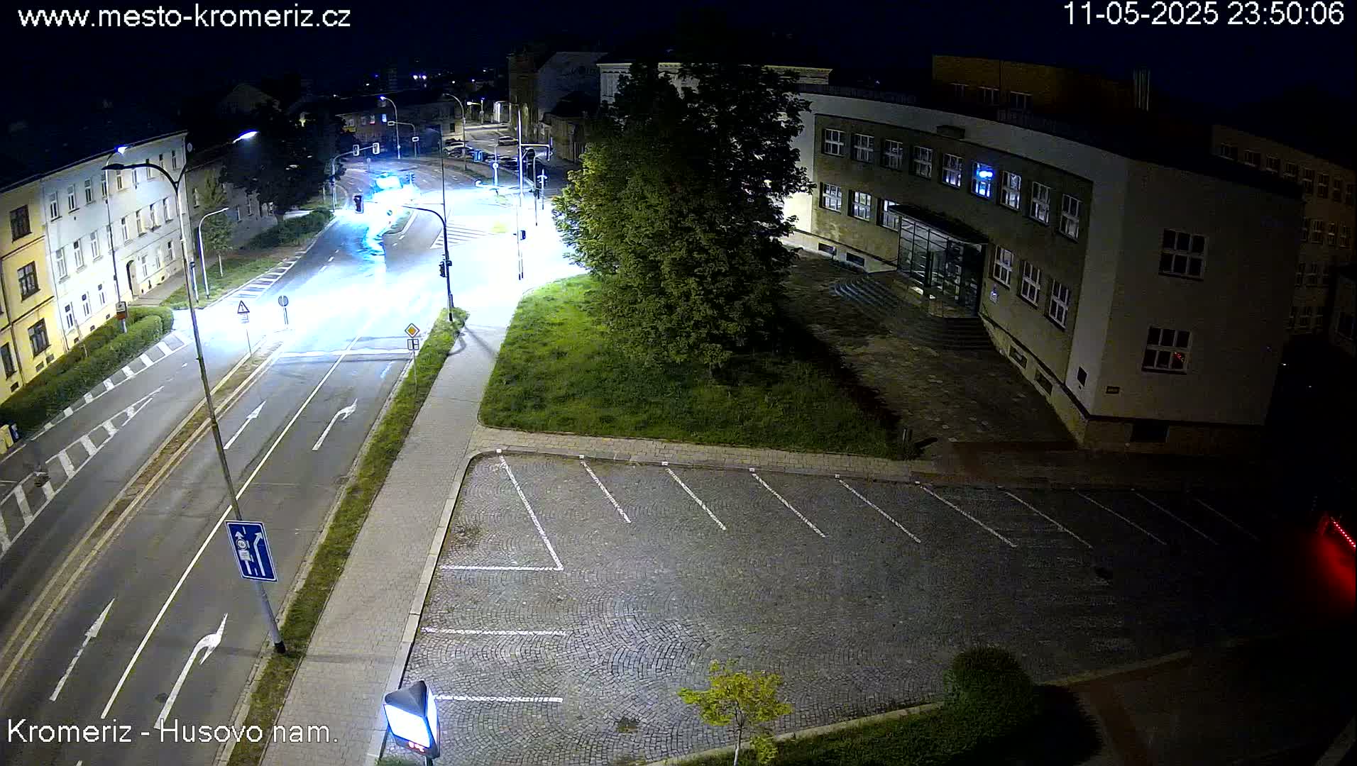 A nighttime, high-angle view shows a paved road with a few cars and a parking lot next to a building, all illuminated by streetlights.