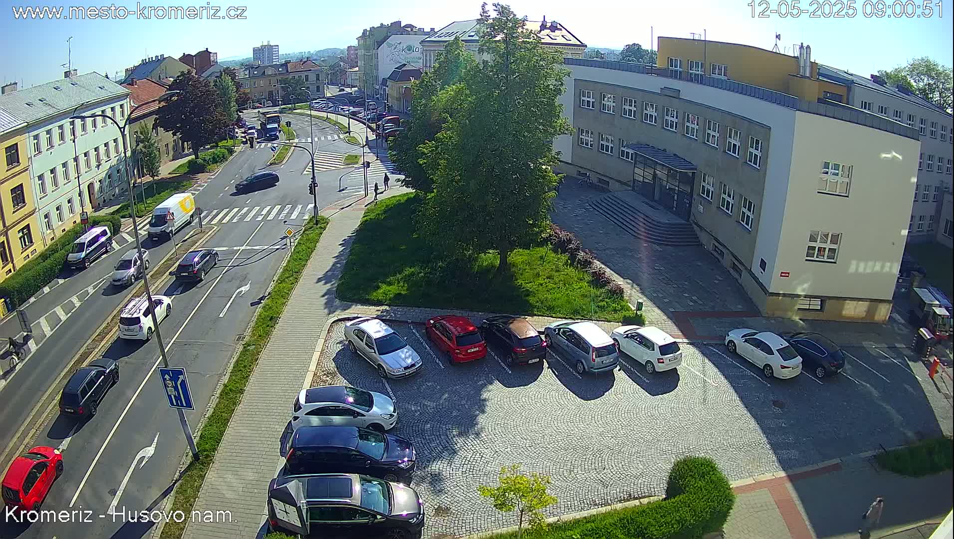 A sunny day view of a town square with cars parked in a cobblestone lot, cars driving on a street, and buildings surrounding a green space.