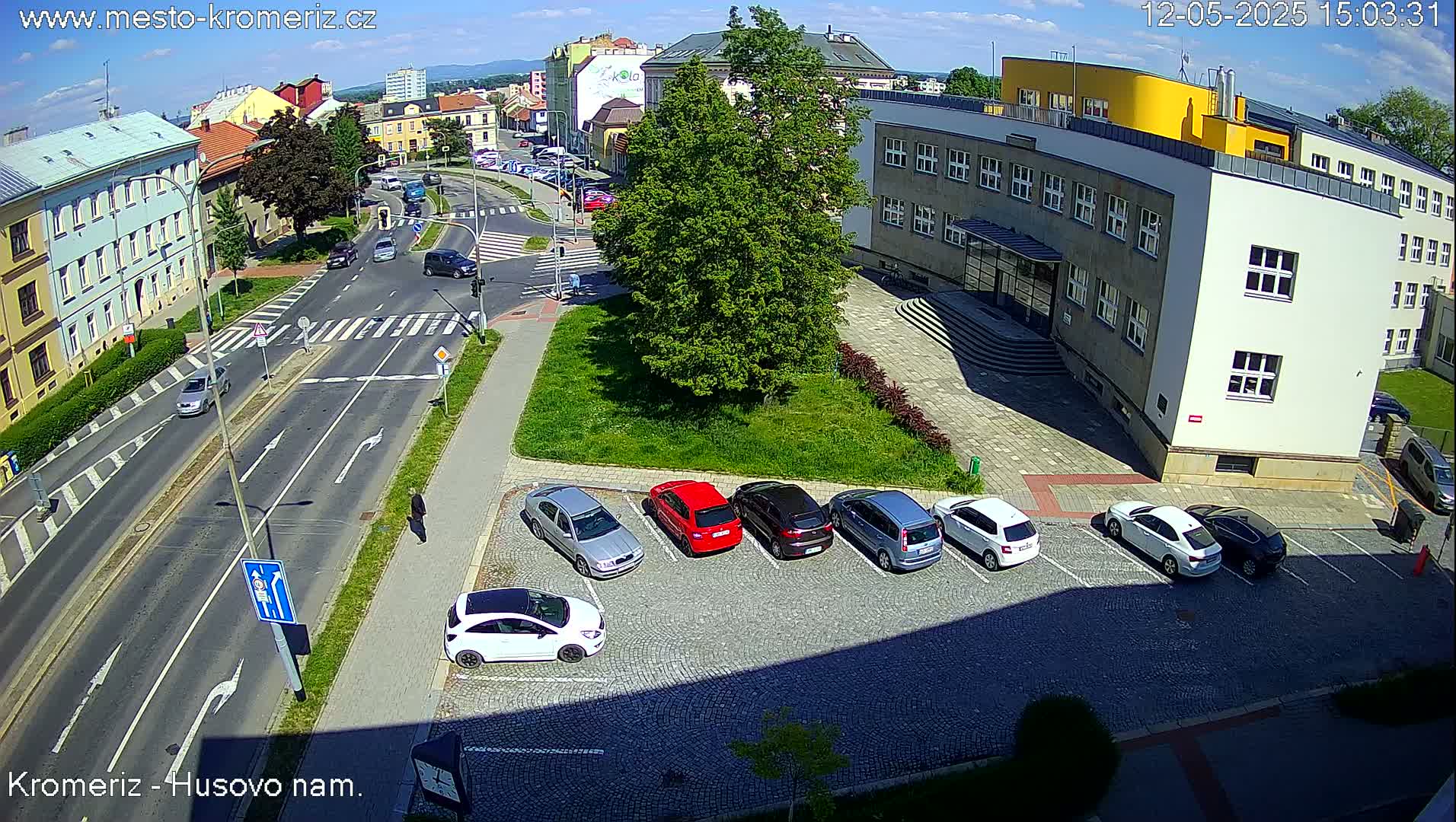A sunny day shows a street intersection with several cars parked and driving,  near a multi-story building.