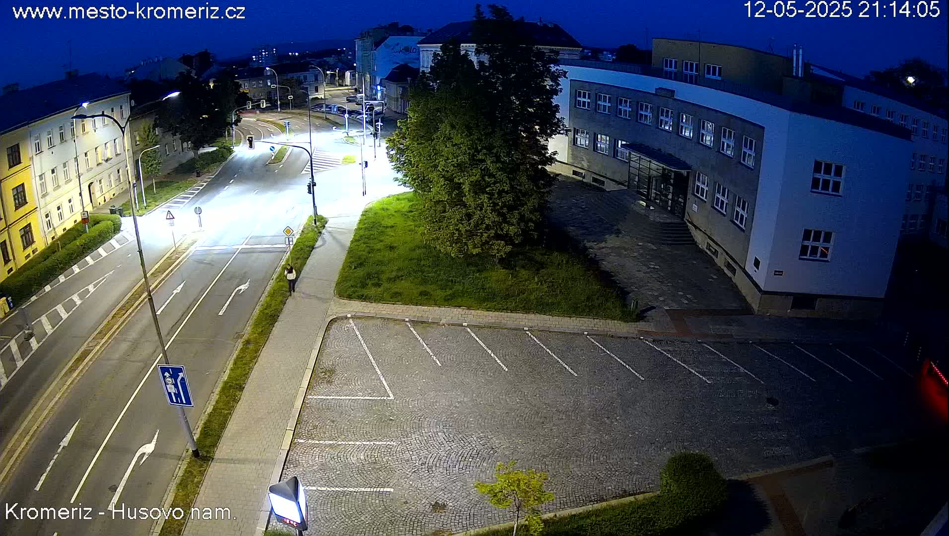 A nighttime, street-level view shows a mostly empty street with a few cars, a pedestrian, and a parking lot next to a building under artificial light.