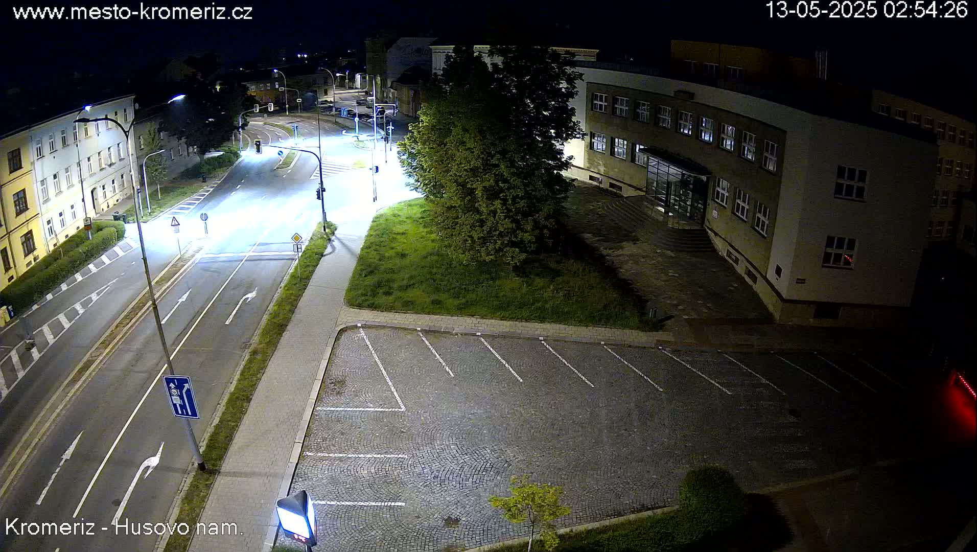 A nighttime, overhead view of an empty paved parking lot next to a multi-lane road and a building with illuminated windows.