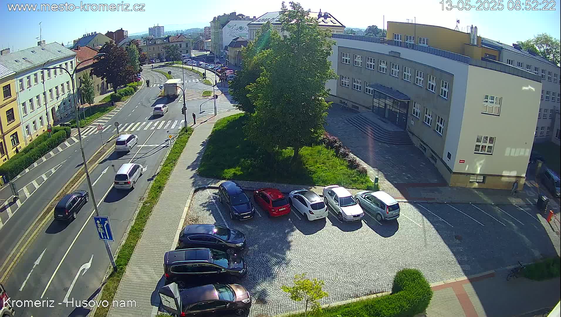 An aerial view shows a sunny city street scene with several cars parked and driving, situated near a large building and green space.