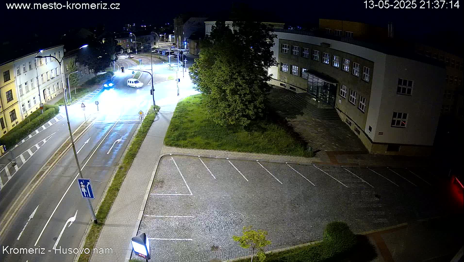 A nighttime, overhead view of a street intersection with a few cars, a parking lot, and a building, illuminated by streetlights.