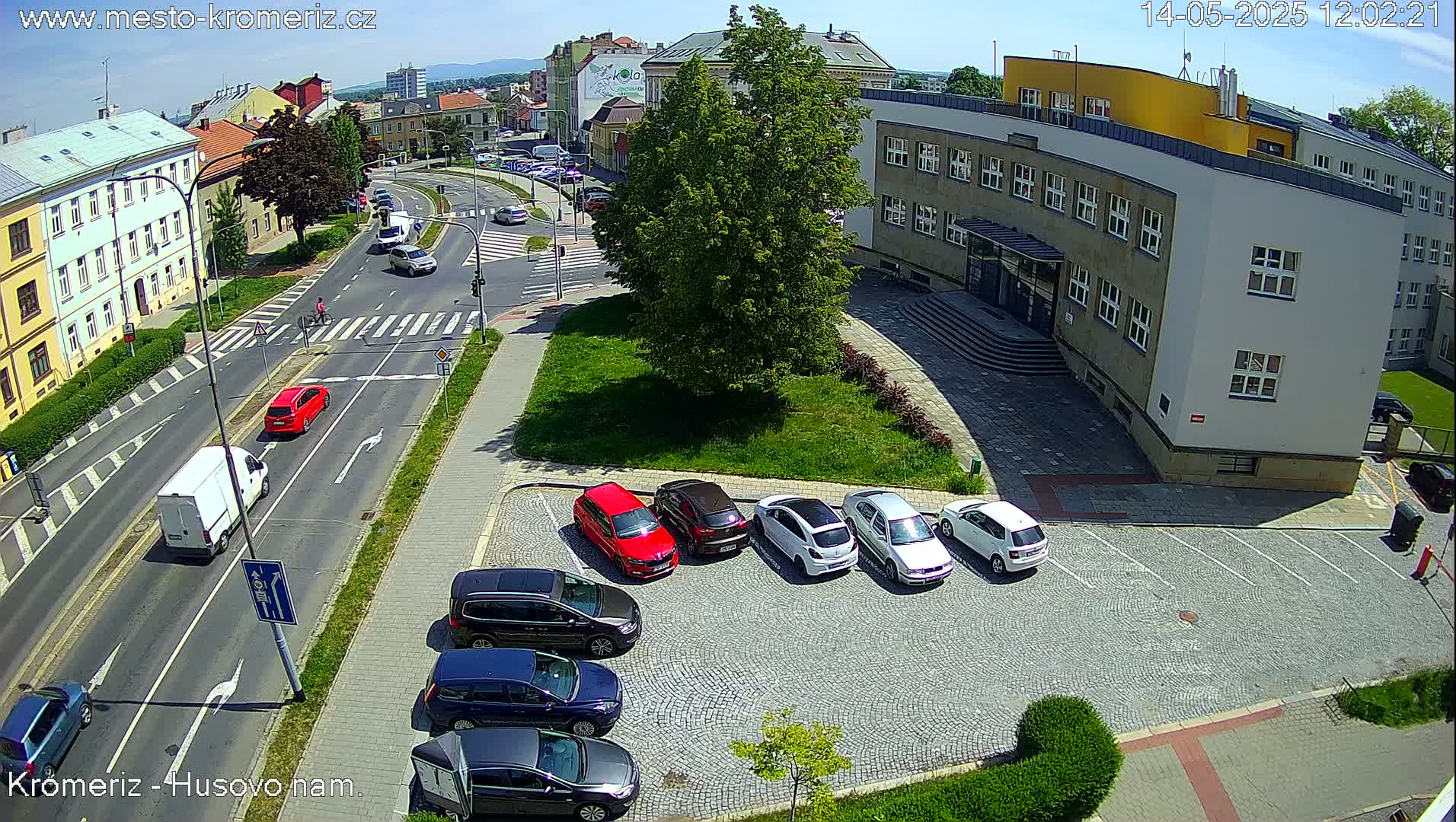 A sunny day shows a street scene with several cars driving and parked, including a parking lot with several cars parked in front of a building.