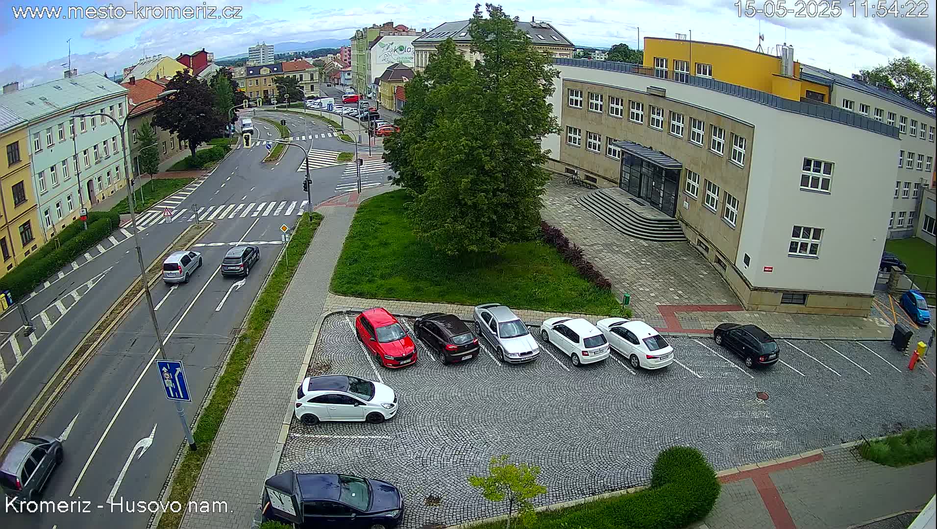 A partly cloudy day reveals a street scene with cars parked in a cobblestone lot next to a multi-story building, and other vehicles driving on a roadway with crosswalks.