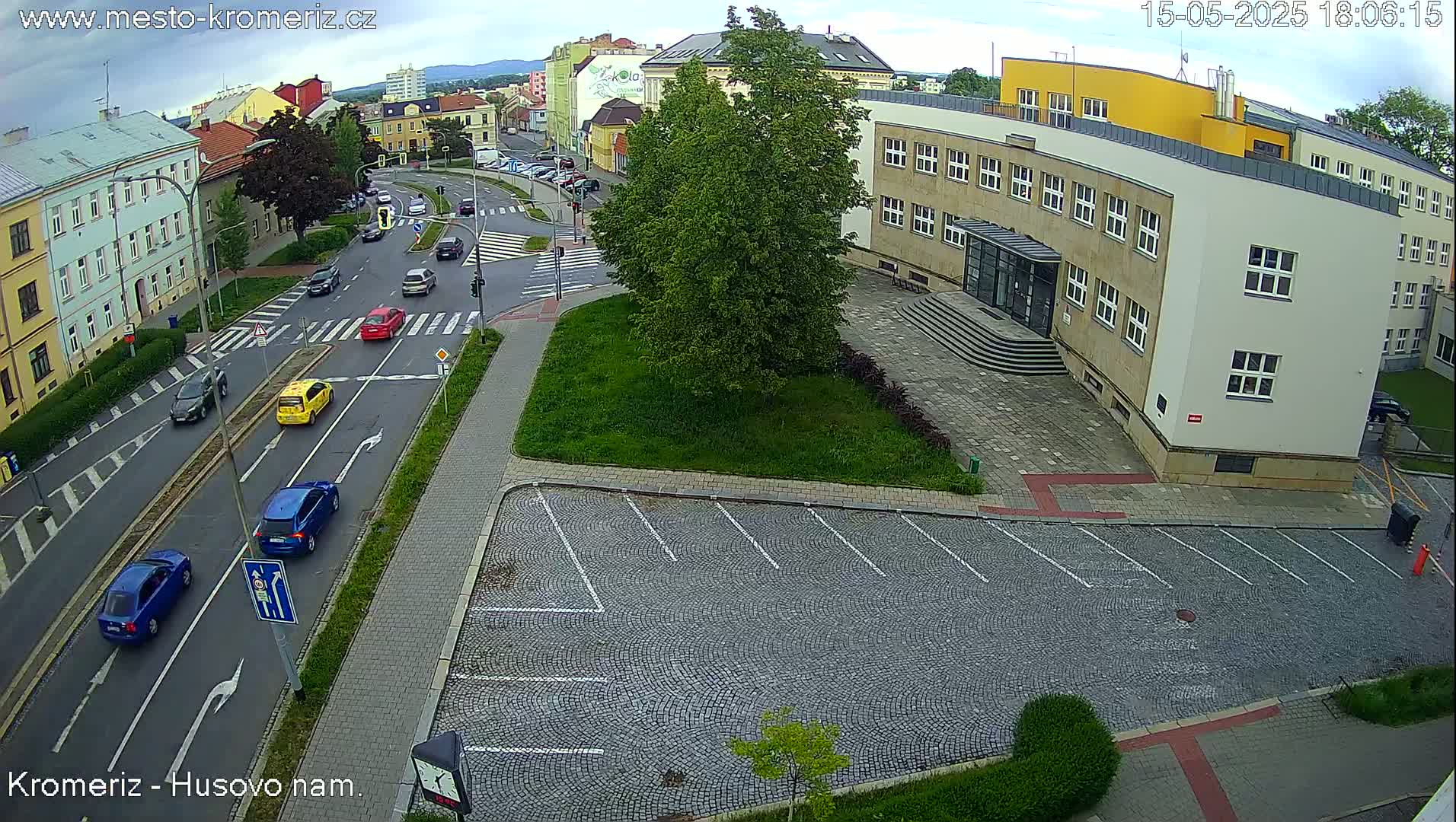 A paved street intersection with several cars, a large building, and an empty cobblestone parking lot, under partly cloudy skies.