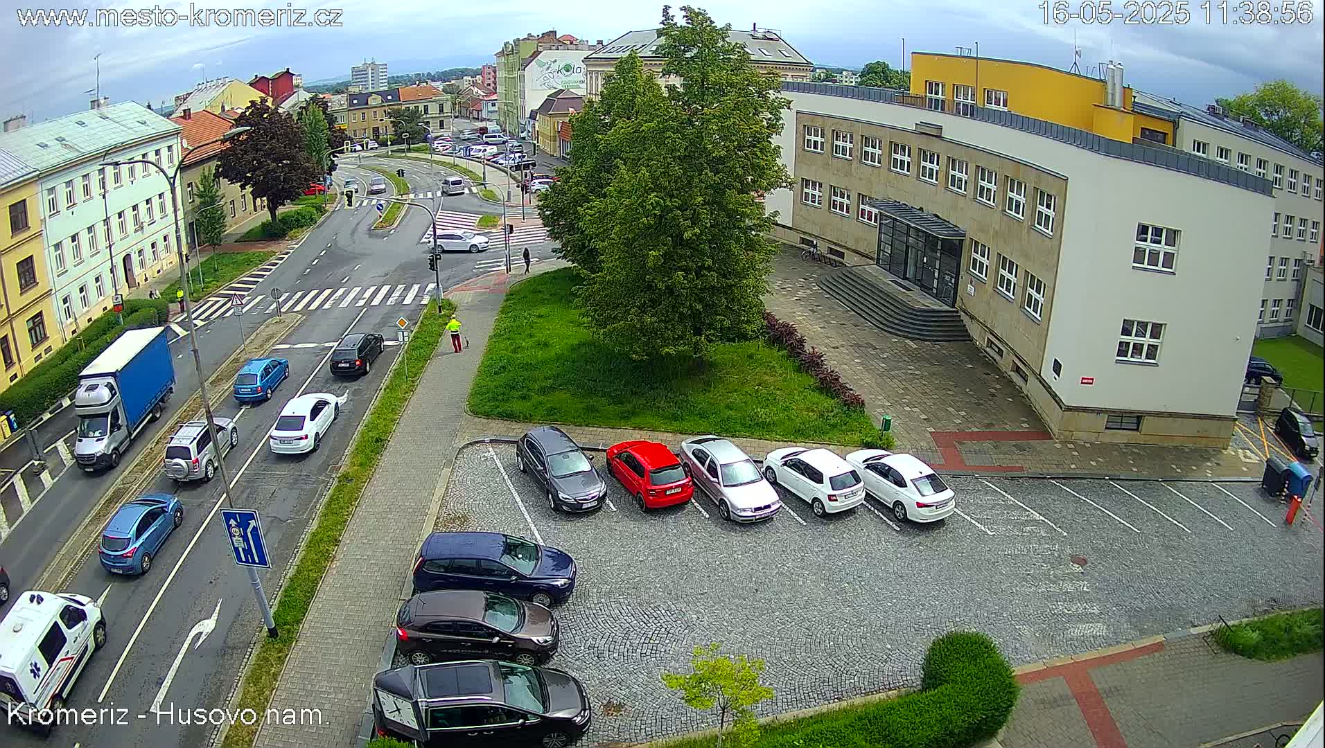 An overhead view shows a city street with several cars, a parking lot with cars parked, and a large building under a cloudy sky.