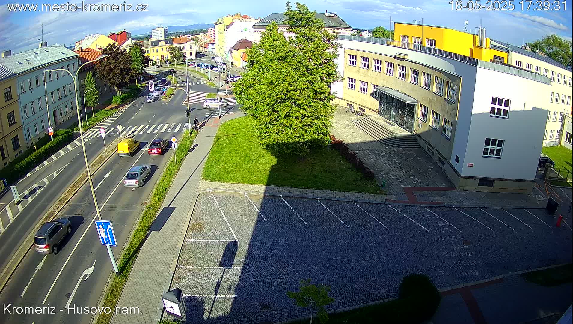 A sunny day shows a street scene with several cars, a large building, a grassy area, and a paved parking lot.