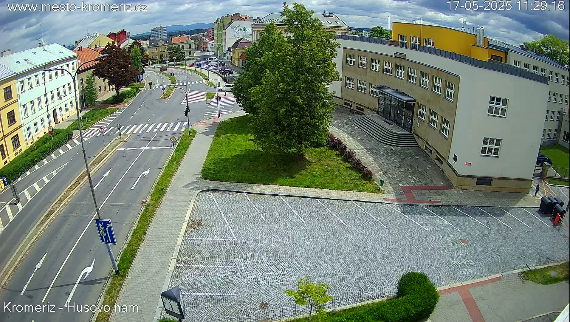 A partly cloudy day reveals a street scene with a large building, a grassy area, and a cobblestone parking lot.