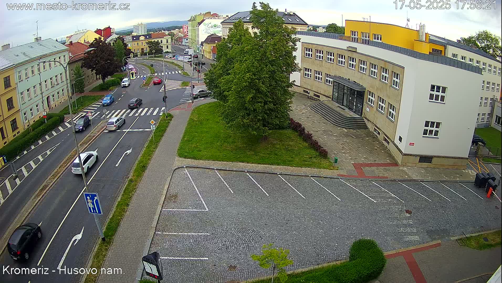 An overcast day reveals a street scene with several cars driving on a road next to a building with an empty cobblestone parking lot.