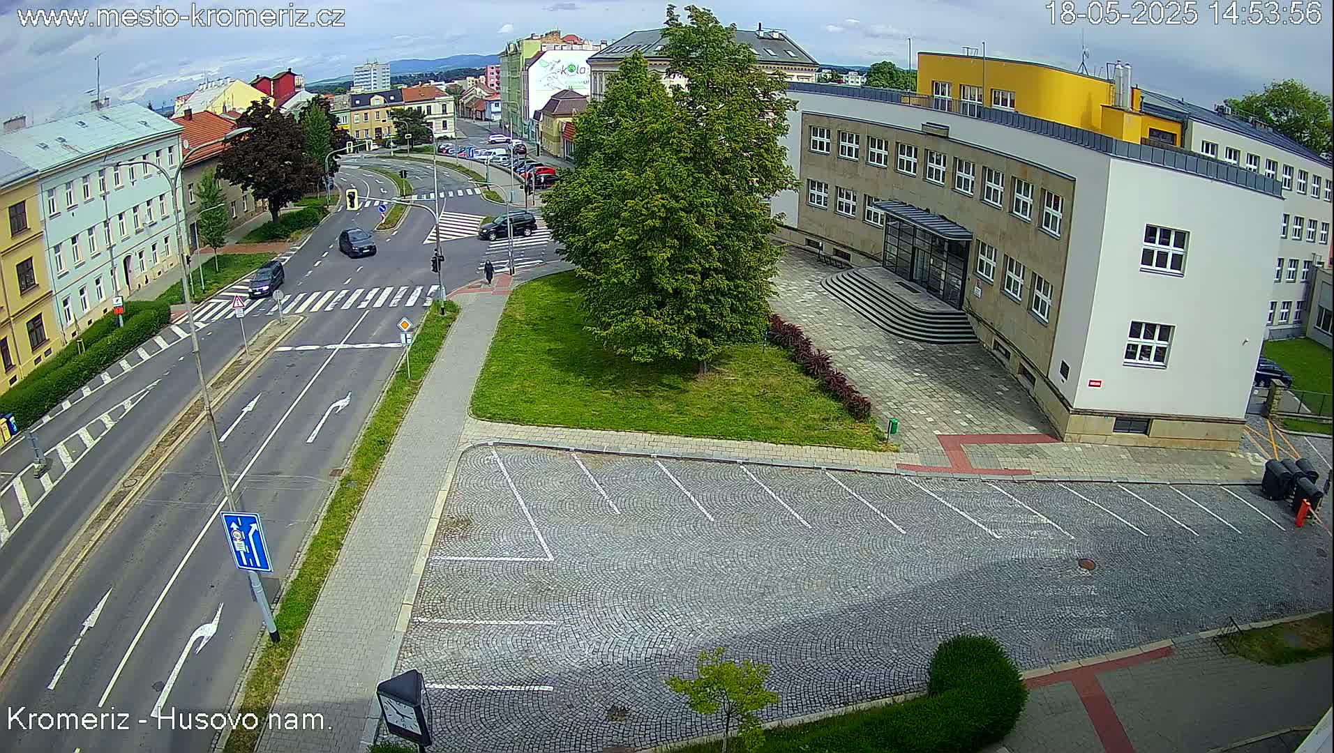 An overhead view of a street intersection and parking lot in a town on a partly cloudy day, showing cars and a large building.