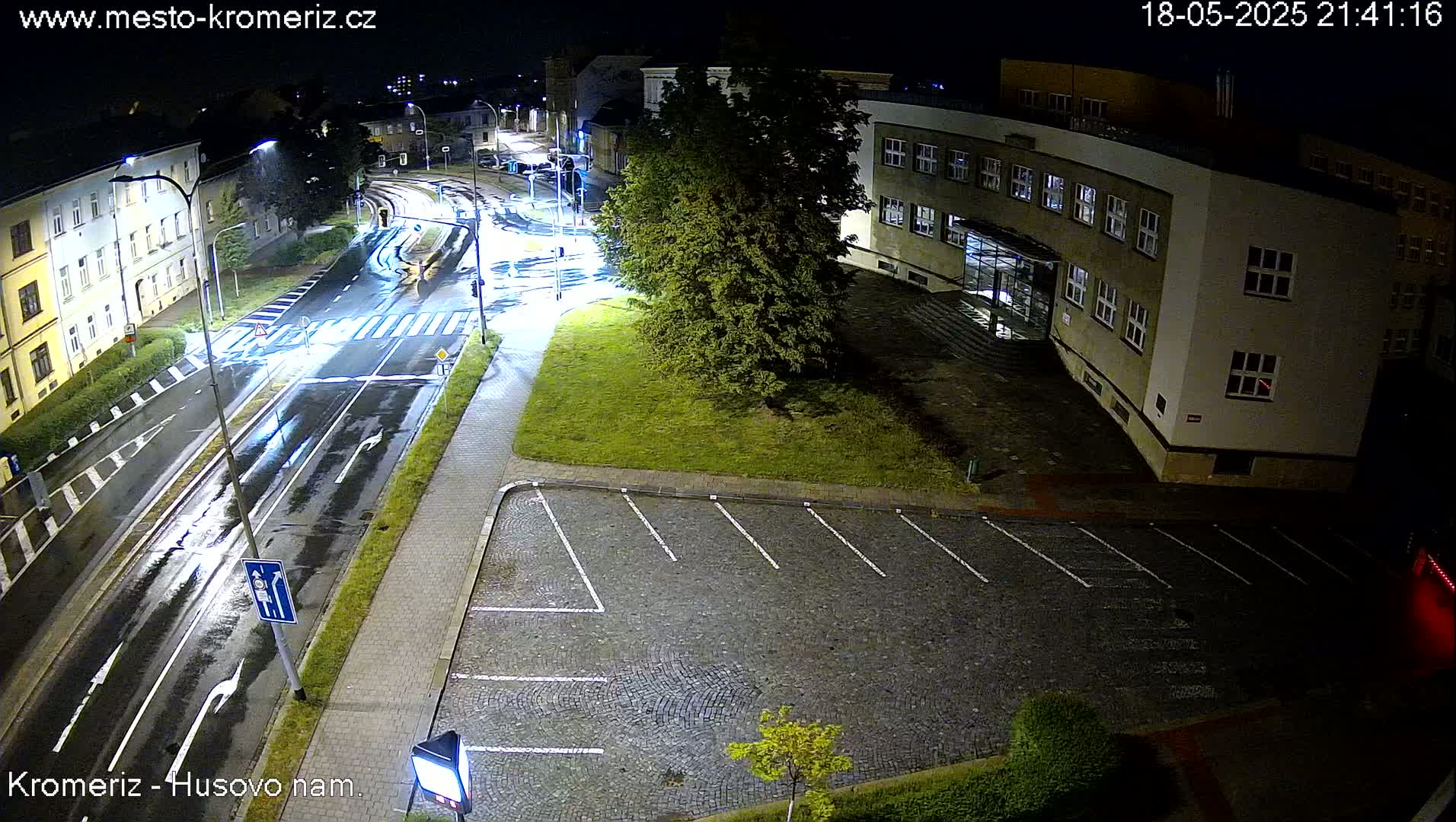A wet, nighttime street scene shows a multi-lane road with crosswalks, a building, a grassy area with a parking lot, and some trees under streetlights.