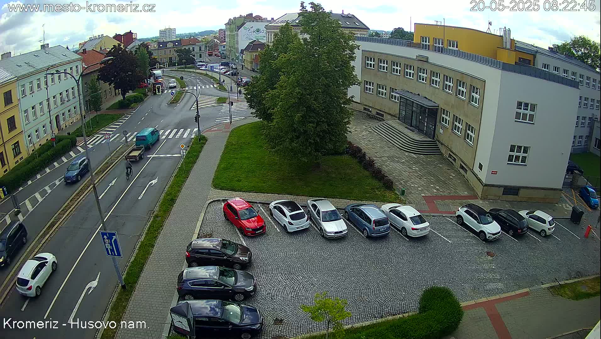 A partly cloudy day shows a street scene with cars parked in a cobblestone lot next to a building and other cars driving on the road.