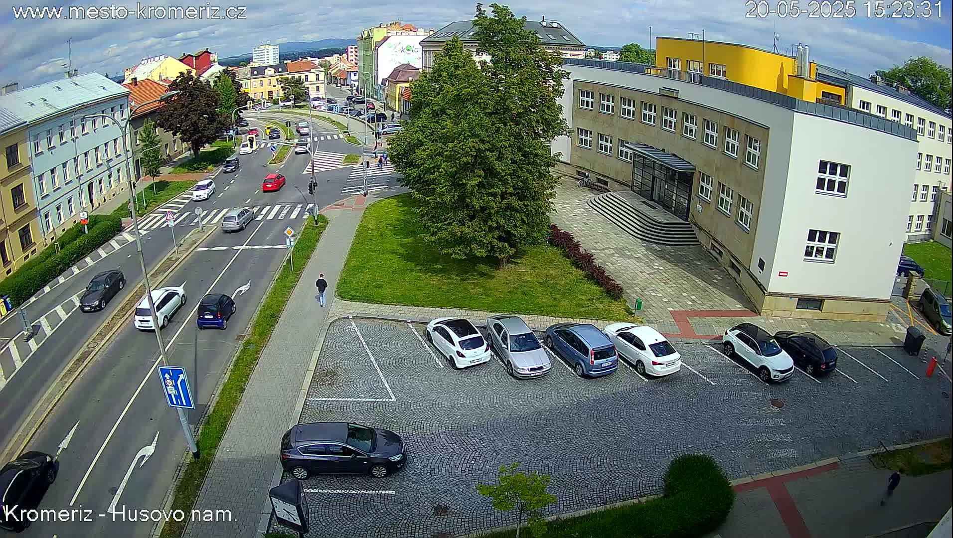 A sunny day shows a street scene with cars parked in a lot next to a building, and cars driving on a road with pedestrians nearby.