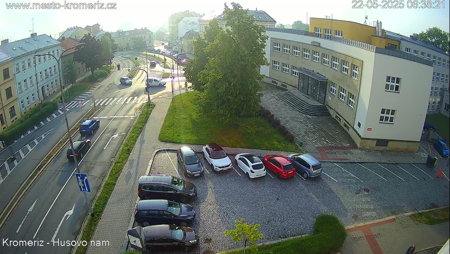 A sunny morning view of a city street with cars parked in a lot next to a building, and several cars driving on the road.