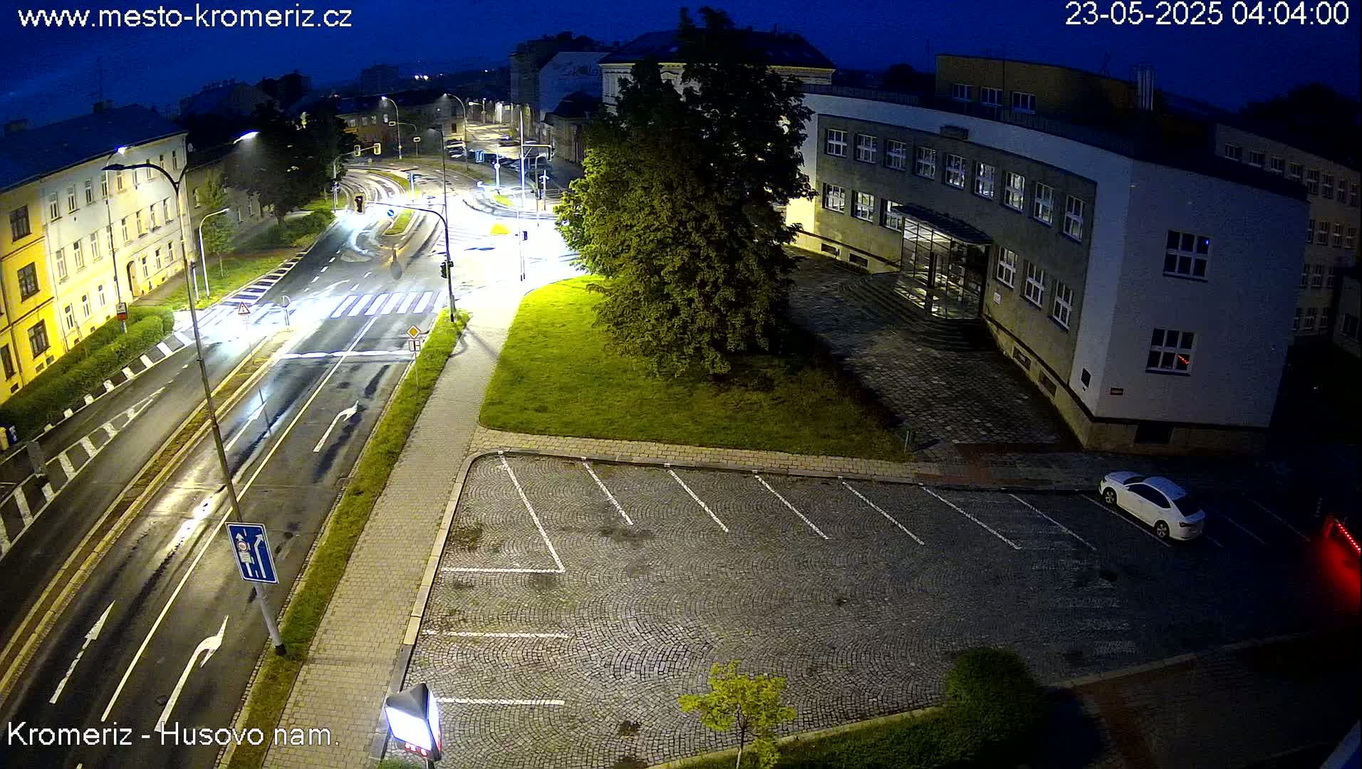 A wet, nighttime street scene shows a multi-lane road with a crosswalk, a building with a parking lot, and a few cars.