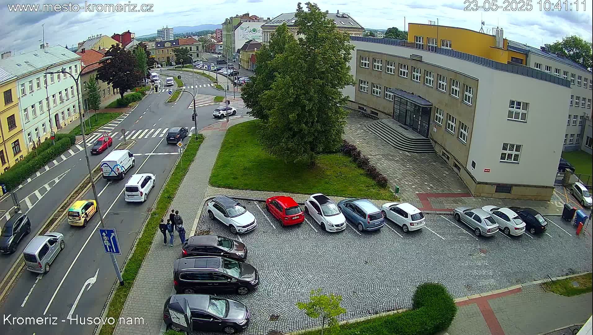 A partly cloudy day shows a street scene with cars parked in a cobblestone lot next to a multi-story building, and other cars driving on roads with pedestrian crossings.