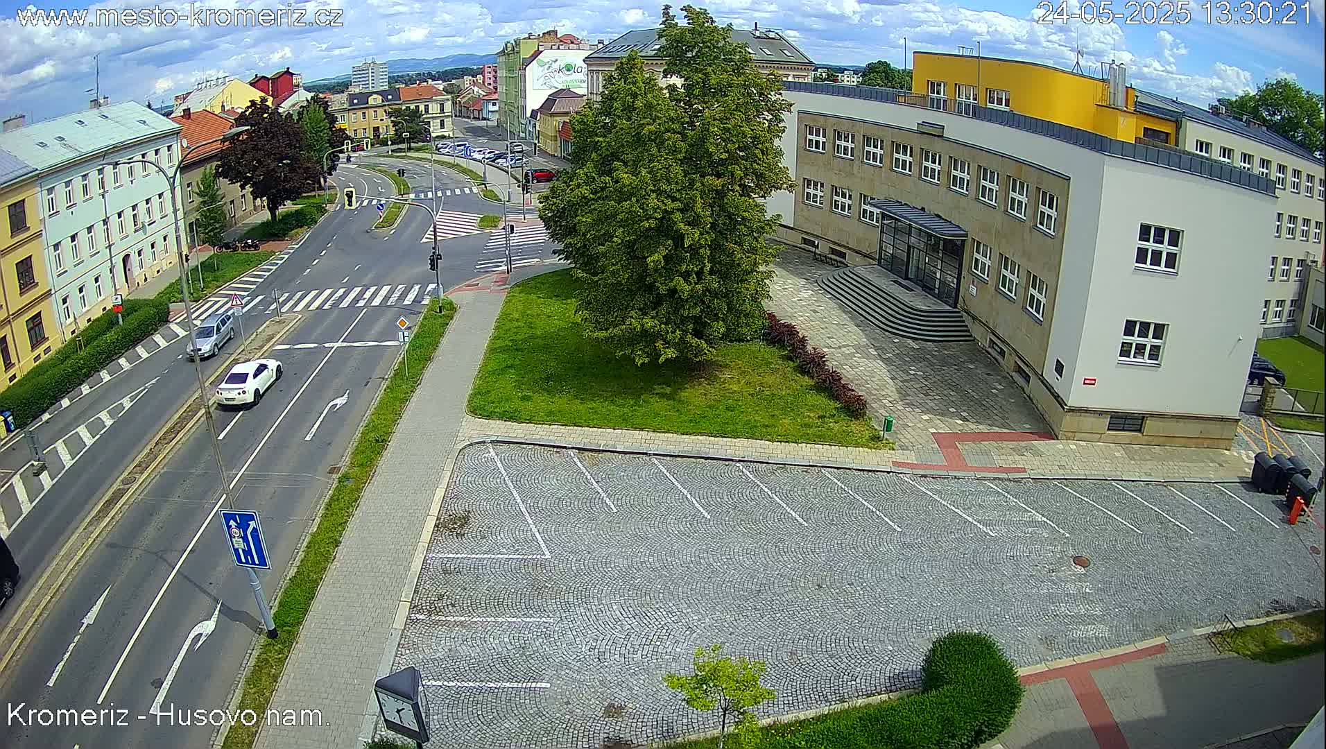 A sunny day reveals a street scene with cars driving on a road, a large building with a parking lot in front, and a small tree in a grassy area.