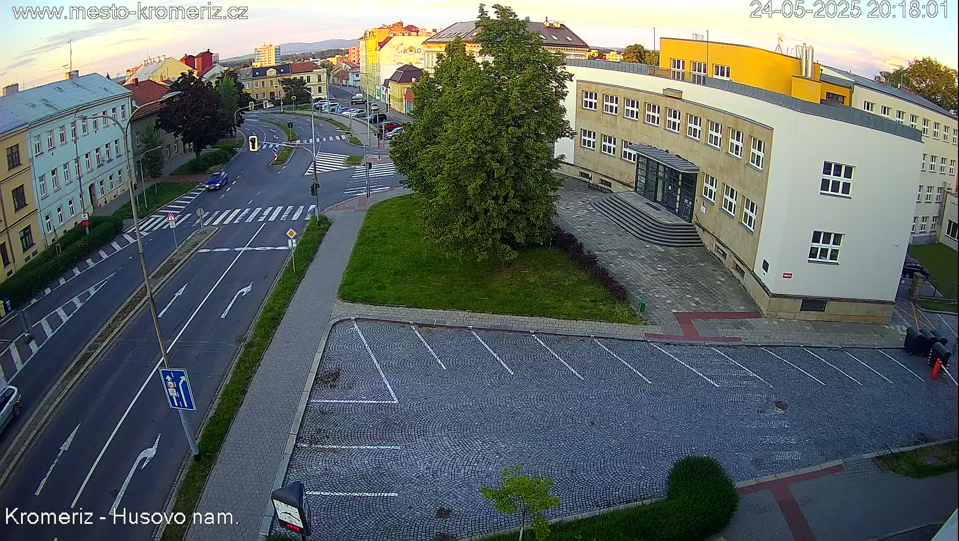An aerial view of a city intersection at dusk, showing a street with cars, a cobblestone parking lot, and a large building.