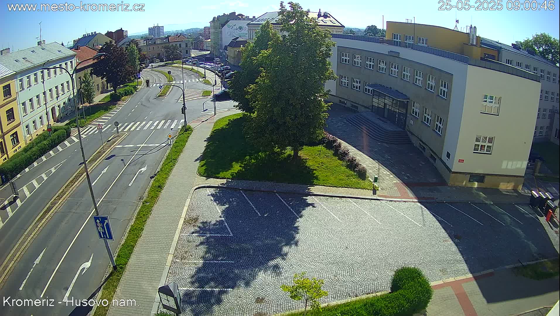 A sunny overhead view of a city street intersection with buildings, a grassy area, and a cobblestone parking lot.