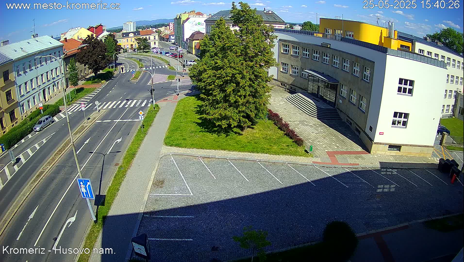 An aerial view shows a sunny city intersection with a large building, a grassy area with a tree, and a paved parking lot.