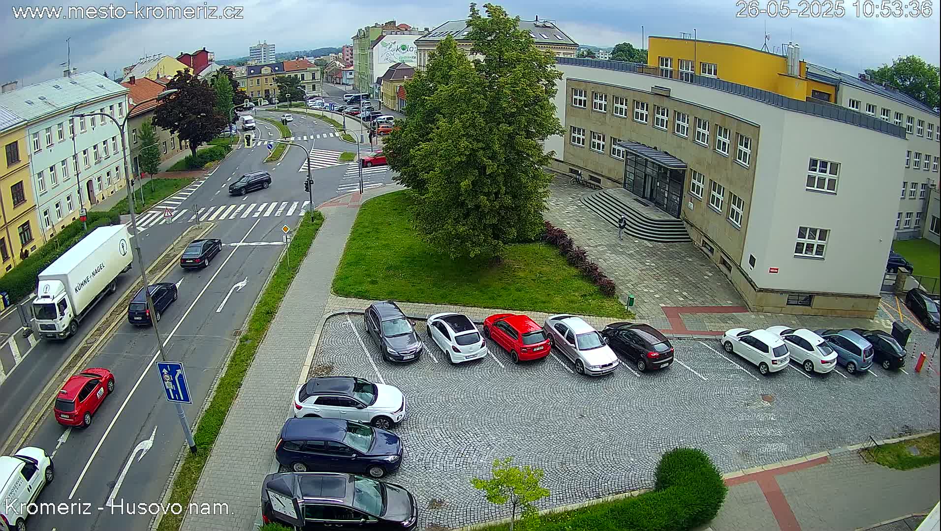 A street scene on a partly cloudy day shows a parking lot with several cars, a street with several cars and a truck, and a large building.