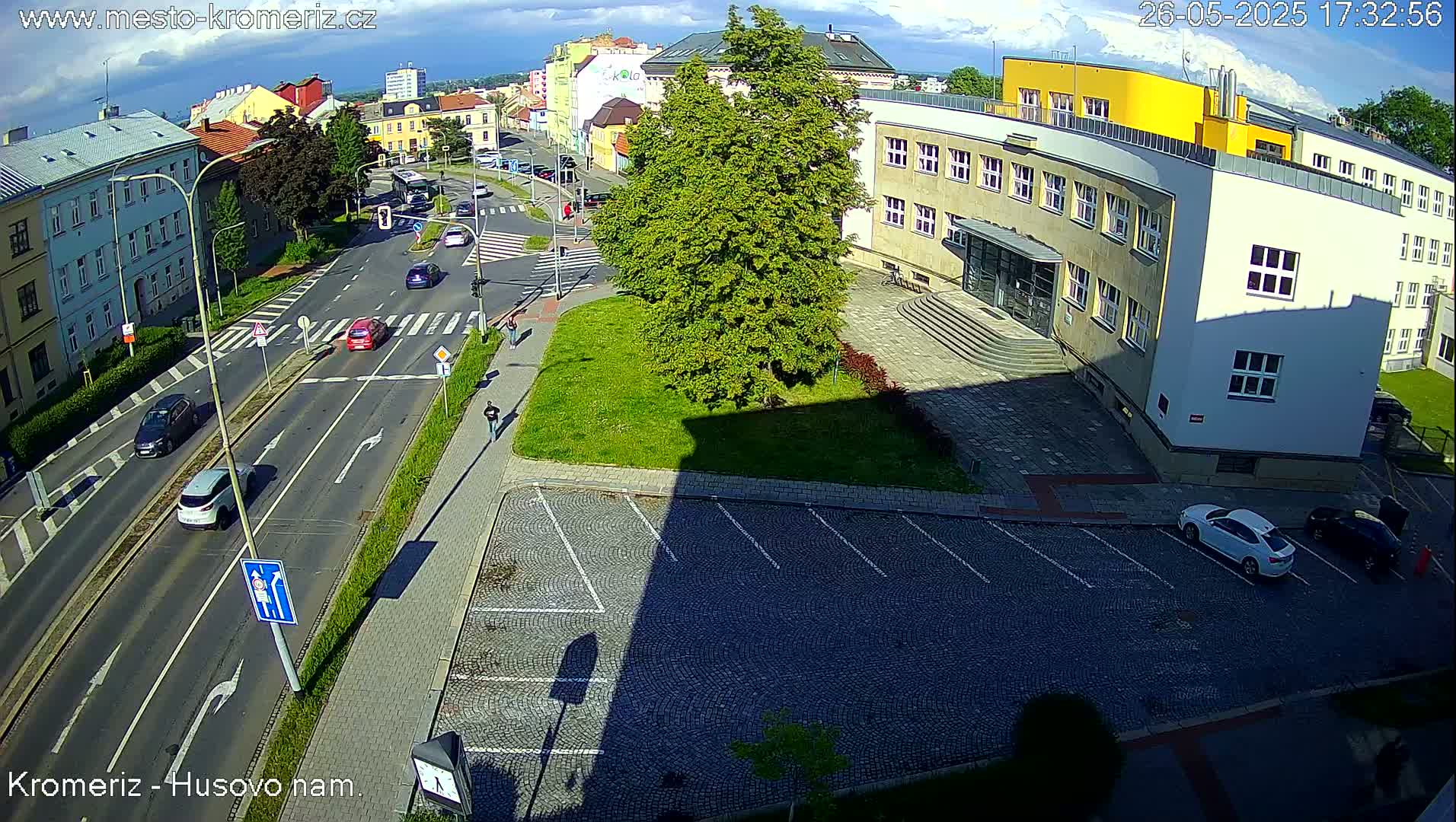 A sunny day shows a high-angle view of a street intersection with several cars, pedestrians, and a large building with a yellow section of the roof.