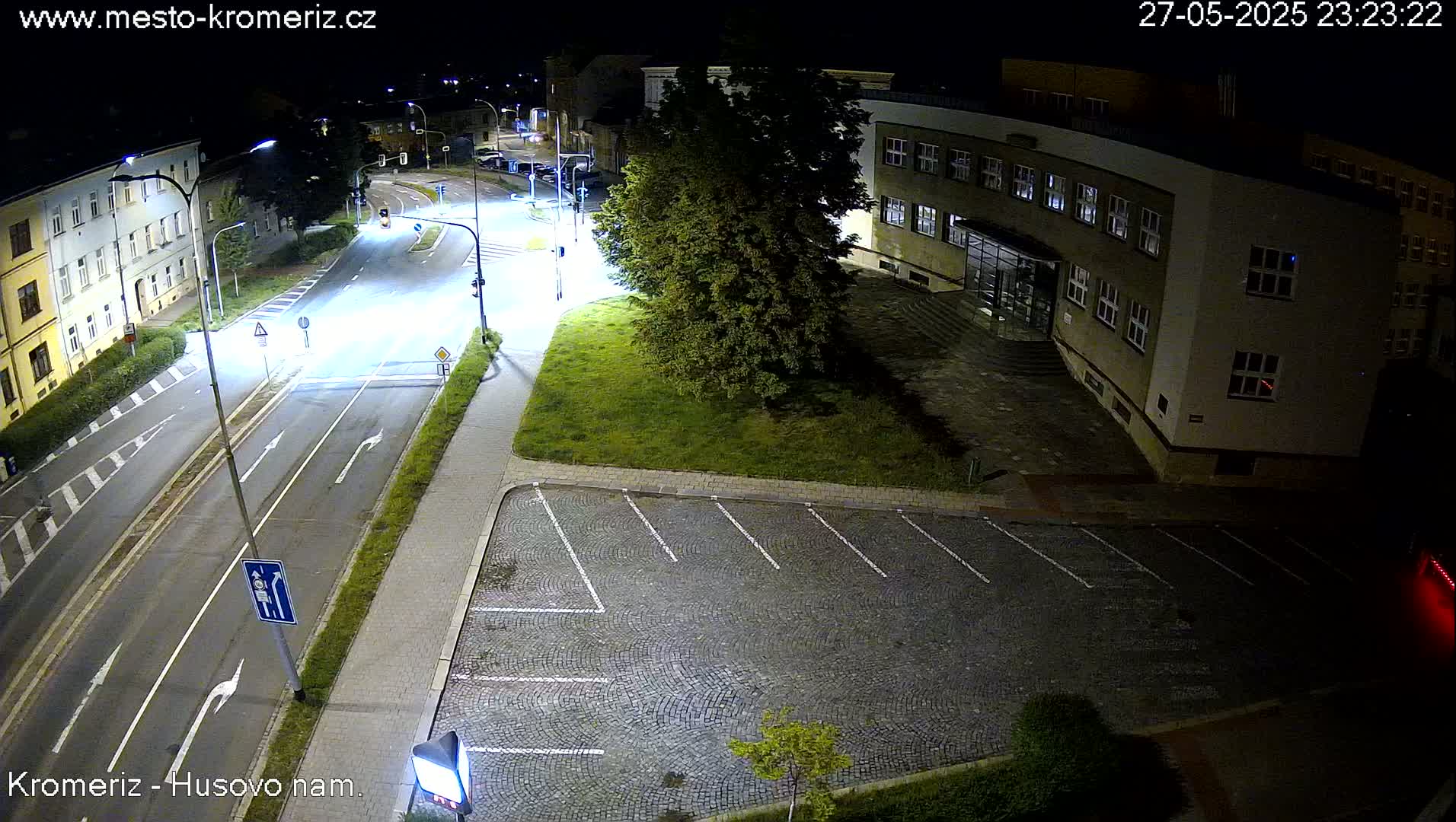 A nighttime, high-angle view of a paved road intersection, an empty cobblestone parking lot, and a building, illuminated by streetlights.