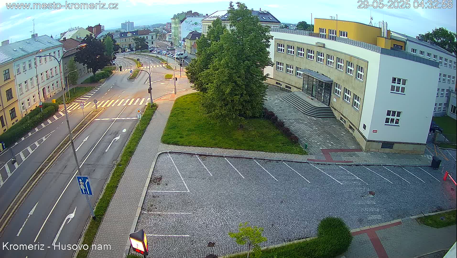 A high-angle, long shot shows a paved road intersection with crosswalks, buildings, a grassy area, and a cobblestone parking lot under a partly cloudy sky.