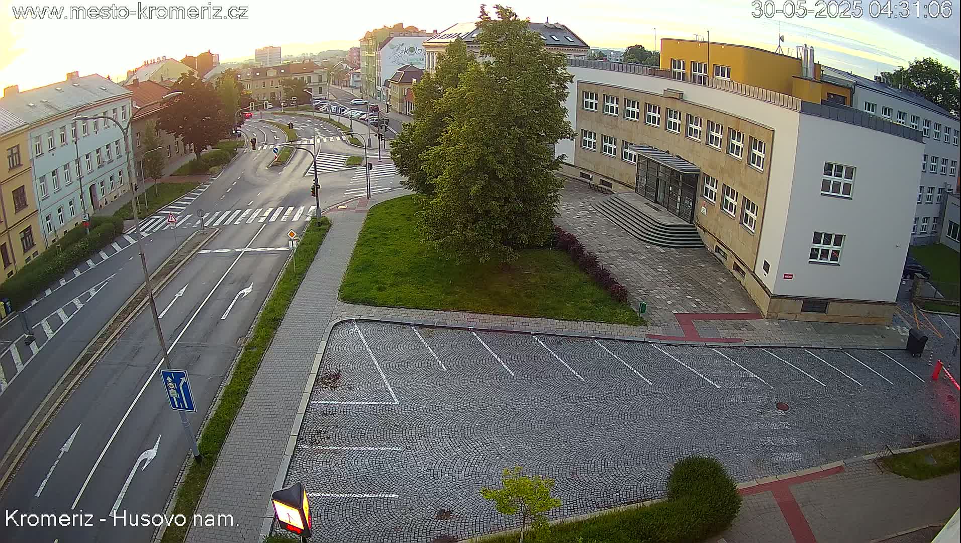 An empty cobblestone parking lot sits adjacent to a multi-story building and a street with light traffic under a partly cloudy sunrise sky.