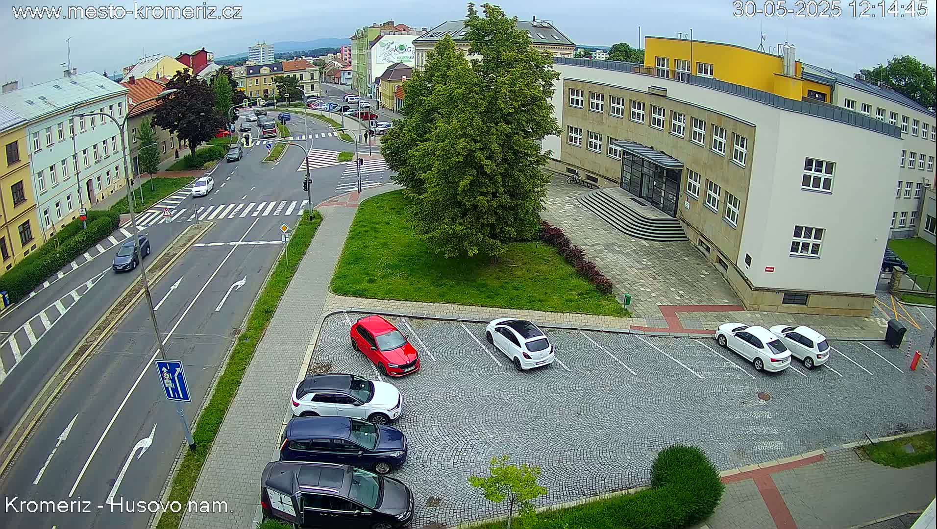 An overcast day reveals a street intersection with several cars parked in a cobblestone lot next to a building, other cars driving on the road, and a large green tree.