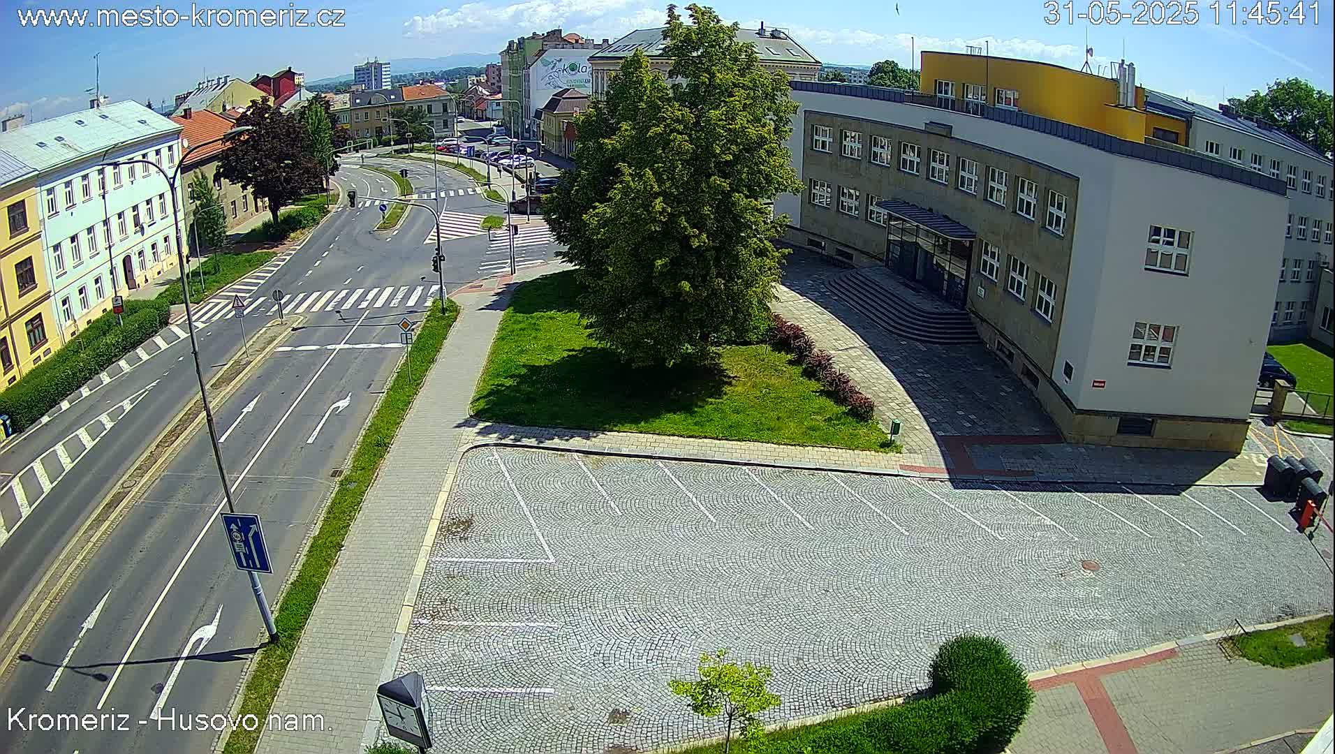 A sunny day overhead reveals a paved roadway intersecting with a cobblestone parking lot next to a large building, with a lush green tree and grassy areas nearby.